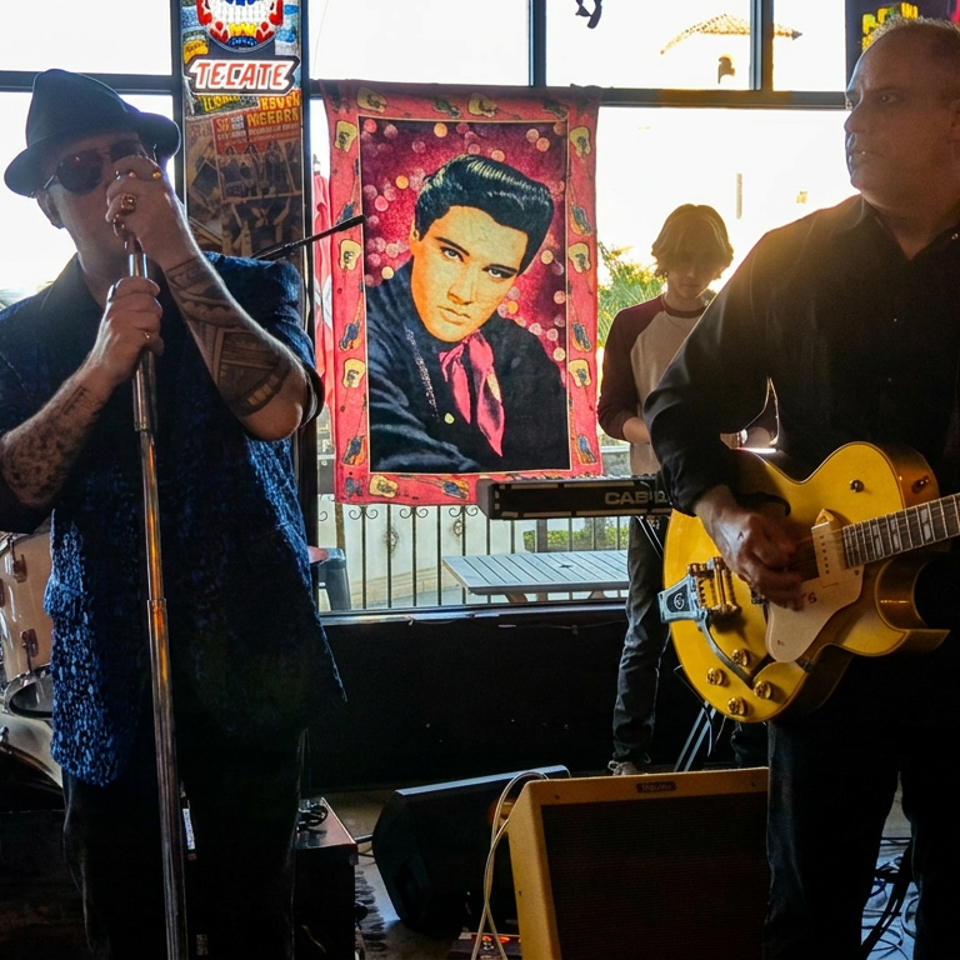 Musicians perform live indoors, with a singer at the microphone, guitarist, drum kit, and colorful rock-and-roll artwork.