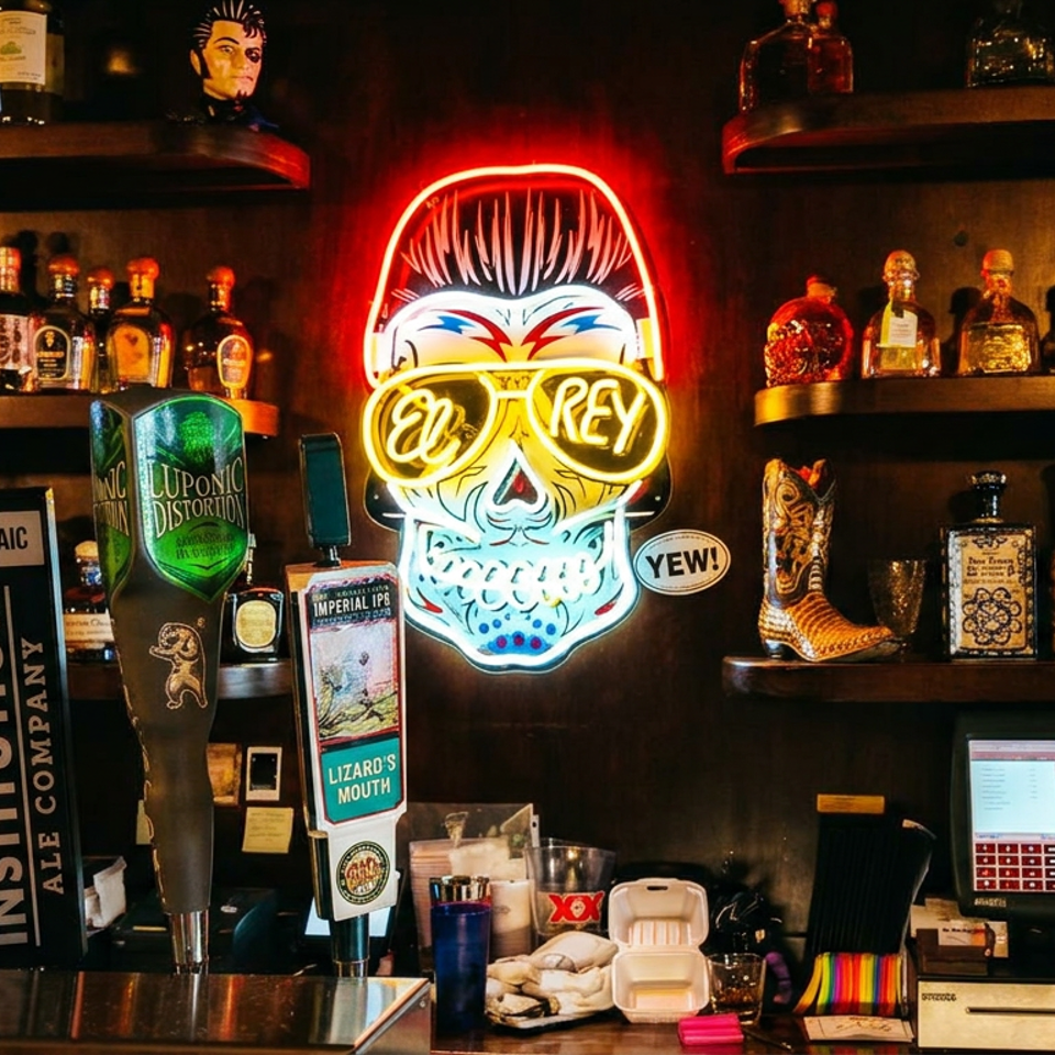 Darkly lit bar counter with beer taps, liquor bottles, colorful El Rey neon sign, decorative skull, and point-of-sale screen.