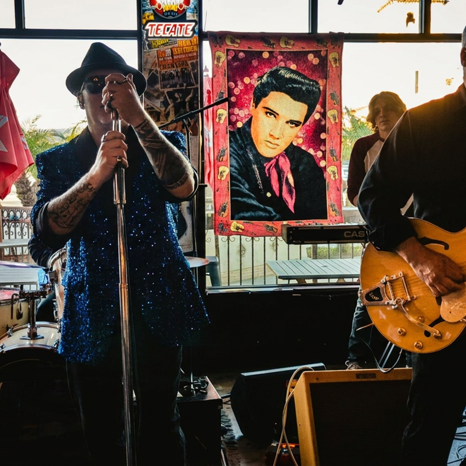 Live band performs indoors, with a singer at the microphone, guitarist, drum kit, and colorful Elvis Presley artwork behind them.